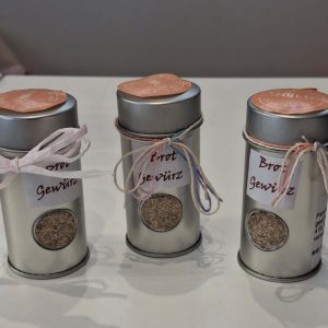 Drei Dosen Brotgewürz mit bunten Bändern und Etiketten auf einem weißen Tisch.
