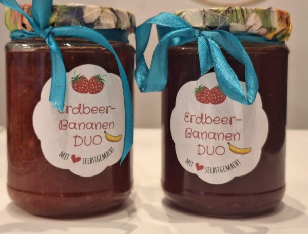 Zwei Gläser Erdbeer-Bananen-Duo-Konfitüre mit bunten Deckeln und blauen Schleifen.