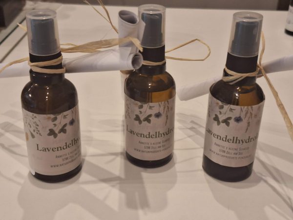 Drei Flaschen Lavendelhydrolat mit dekorativem Etikett und Papierrolle am Tisch.